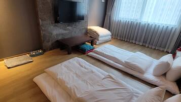 A modern hotel room with a flat-screen TV mounted on a textured wall, a wooden floor, a sofa bed with white bedding, a small table with a remote control, and a window with sheer curtains.