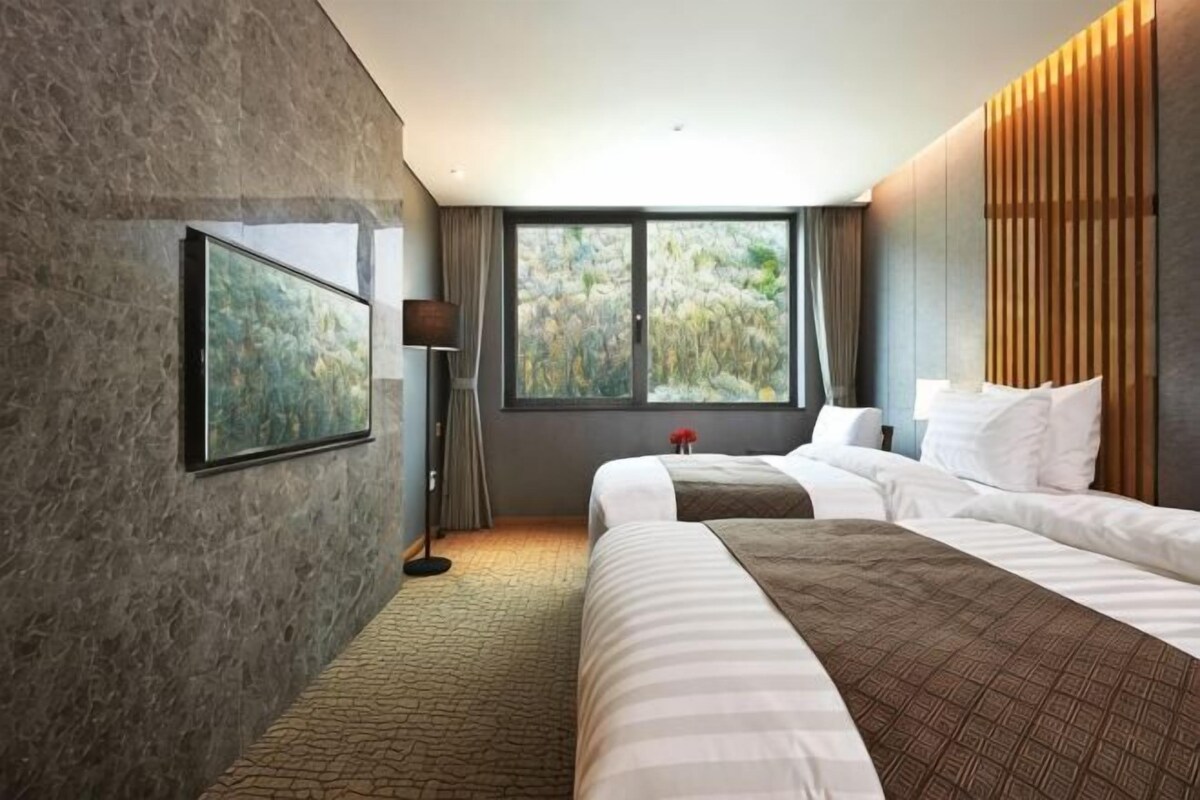 Habitación de hotel con una cama grande, una mesita de noche, una lámpara y una ventana con vista a árboles.