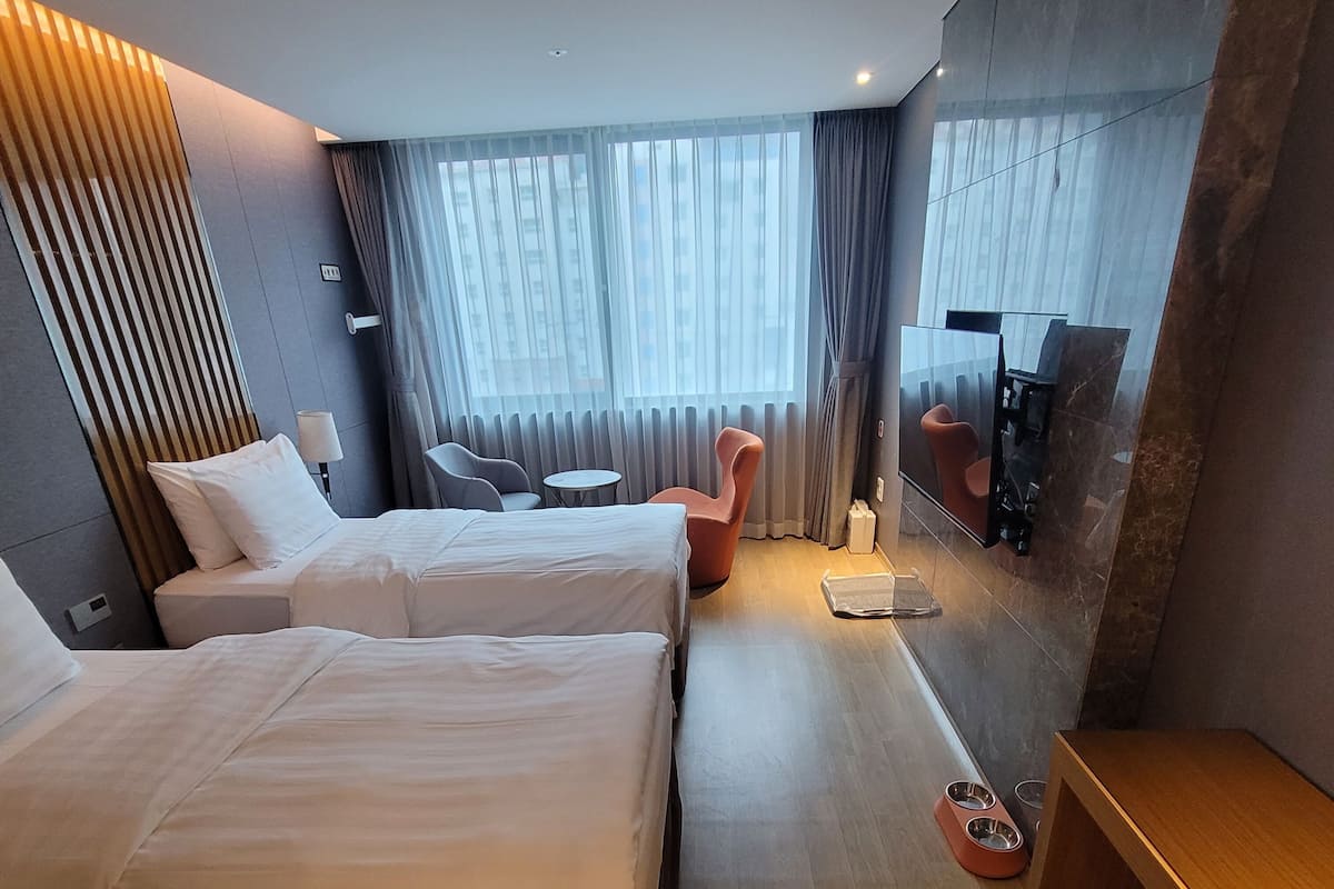 A hotel room with two beds, a small table, a chair, a TV, and a large window with curtains.
