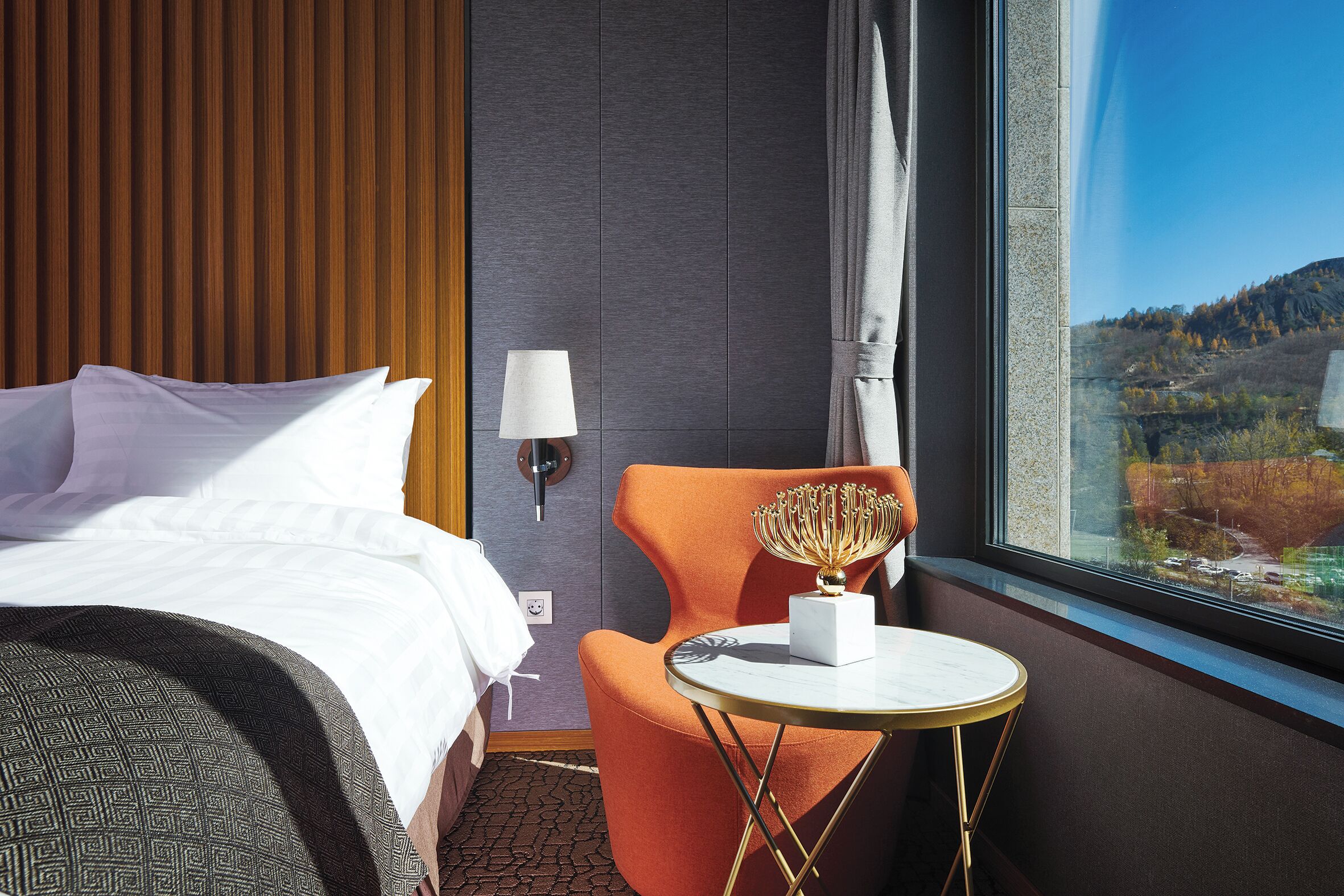 A hotel room with a bed, an orange chair, a small table, and a view of a landscape through a window.