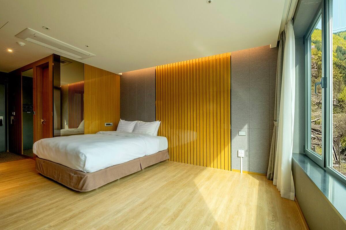 A modern bedroom with a large bed, wooden flooring, a window with curtains, and a view of trees outside.