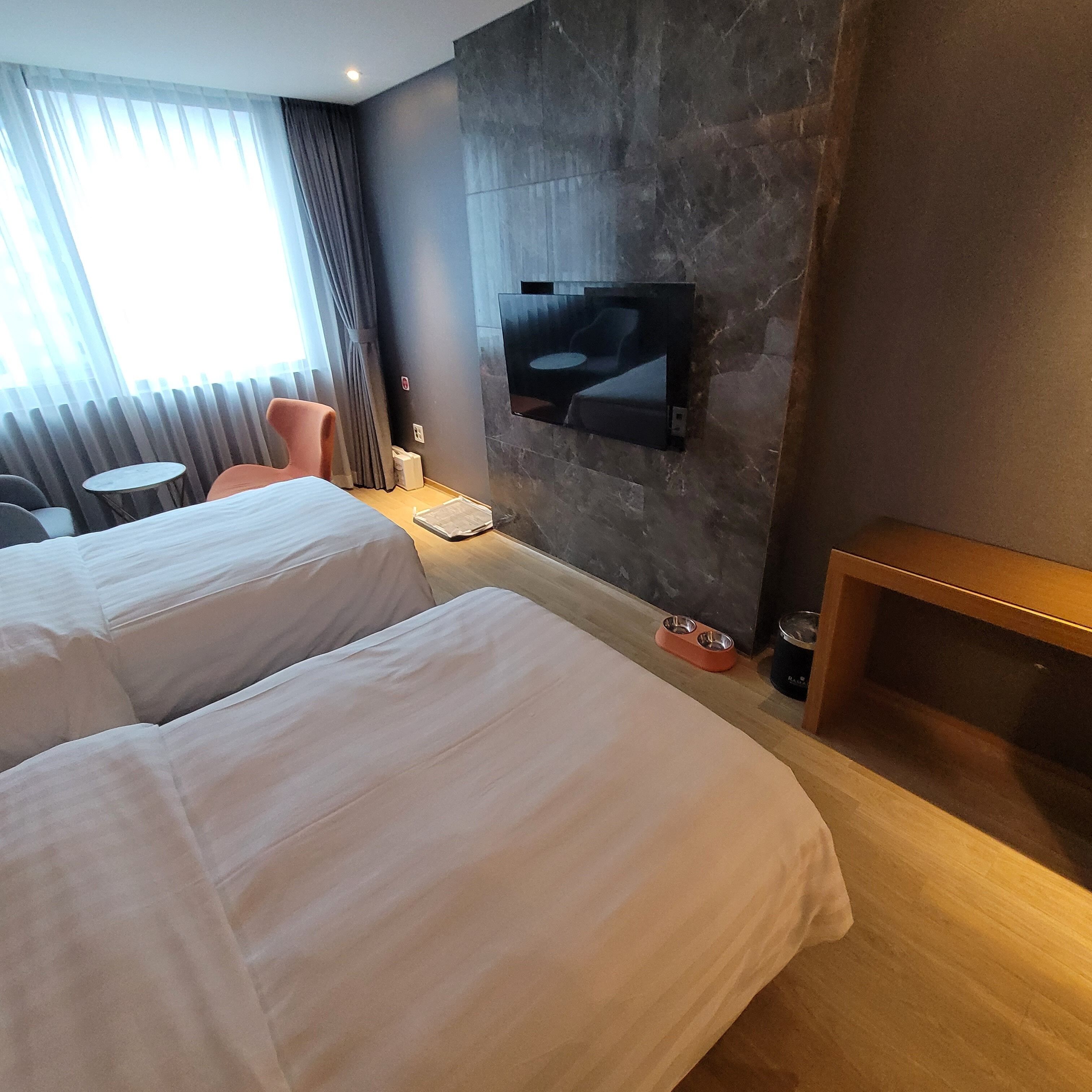 Quarto de hotel com duas camas, uma televisão presa à parede, uma mesinha e uma cadeira.