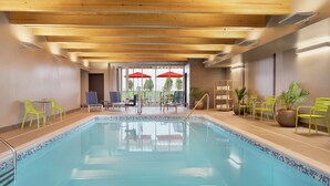 Una piscina interior con vigas de madera en el techo, un borde de azulejos y varias sillas y sombrillas afuera.