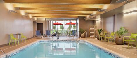 An indoor swimming pool with wooden beams on the ceiling, a tiled border, and several chairs and umbrellas outside.