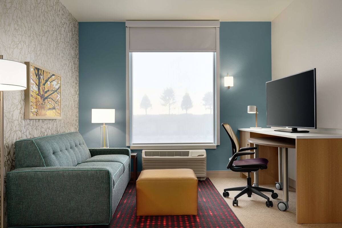 Ein Hotelzimmer mit Sofa, Hocker, Schreibtisch, Stuhl, Fernseher, Fenster, Klimaanlage und Lampen.