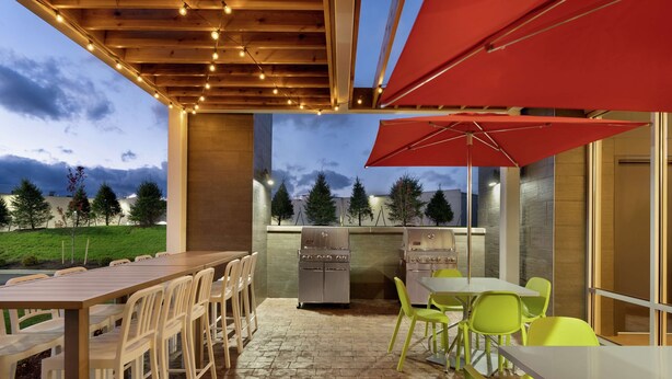 Un patio al aire libre con una larga mesa de madera y sillas, un paraguas rojo y una estación de parrillada.
