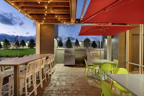 An outdoor patio area with a long wooden table and chairs, a red umbrella, and a grill station. - Home2 Suites by Hilton Martinsburg, WV (Martinsburg)