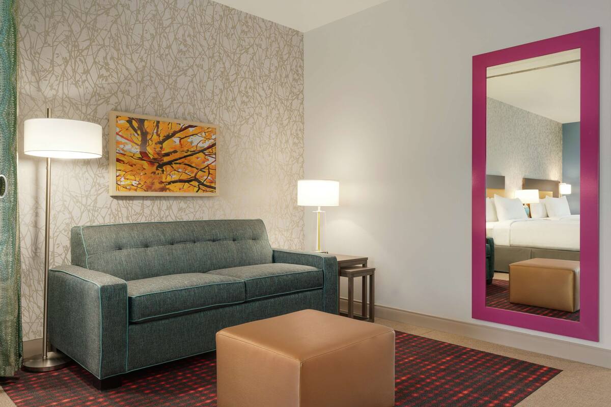 Ein Hotelzimmer mit einem blaugrünen Sofa, einem kleinen Hocker, einer Stehlampe, einem Tisch und einem großen Spiegel mit rosafarbenem Rahmen.