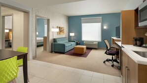 Una habitación de hotel moderna con sofá, reposapiés, mesa de comedor, sillas, kitchenette y un ventanal grande.