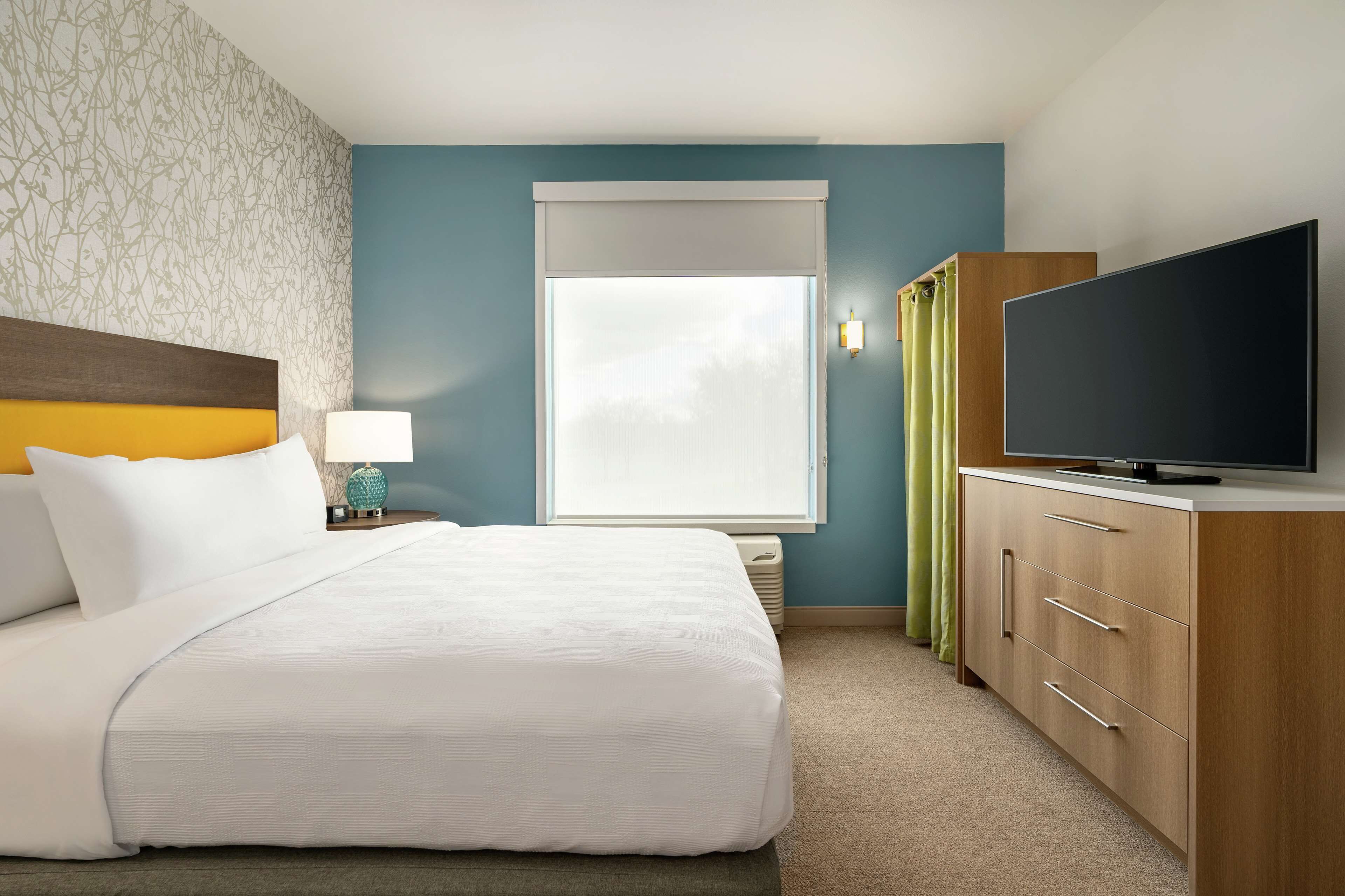 A hotel room with a bed, a TV, a nightstand, a lamp, a window with curtains, and a wardrobe.