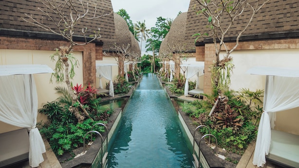 A corridor with a central water channel flanked by two buildings with thatched roofs, surrounded by lush greenery and white canopies. - Tanamas Villas Ubud (Mas)