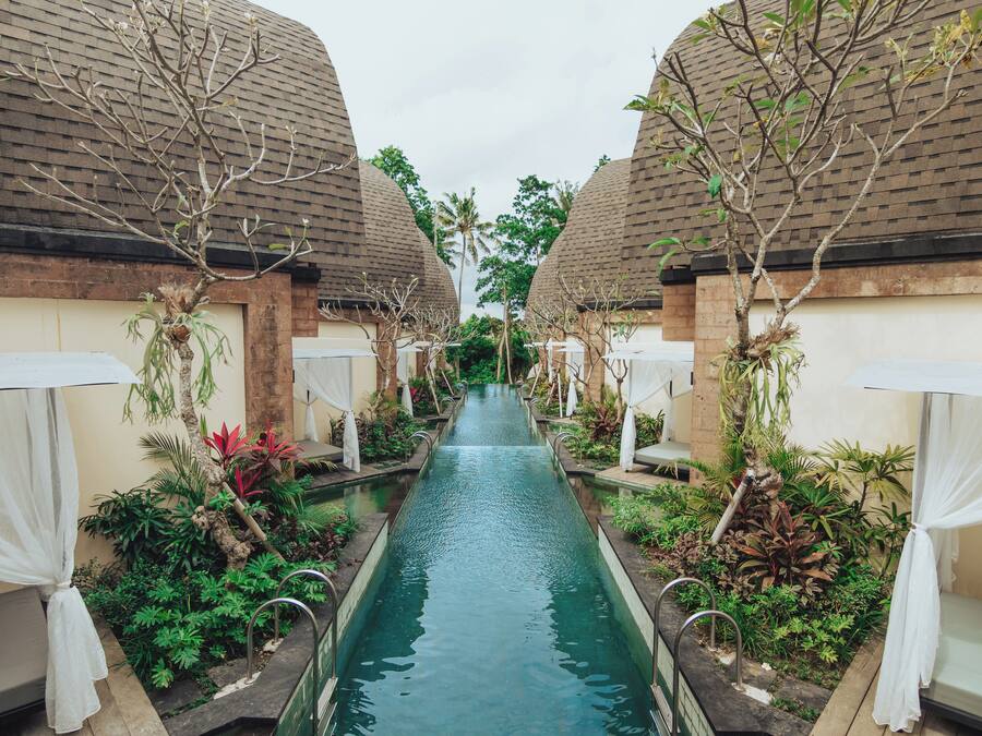 A corridor with a central water channel flanked by two buildings with thatched roofs, surrounded by lush greenery and white canopies.