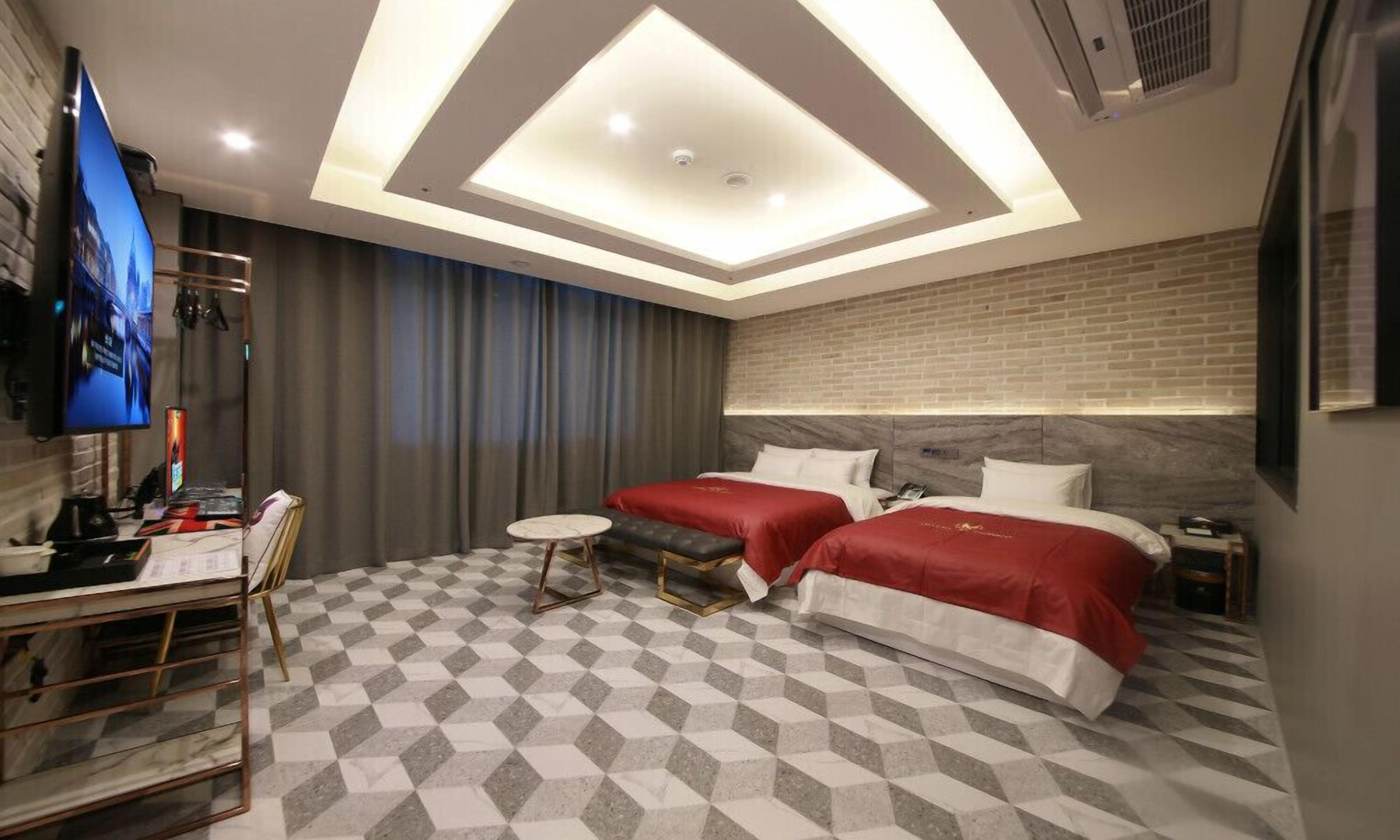 Une chambre d’hôtel moderne avec un sol à carreaux, deux lits avec une literie rouge, un bureau avec un téléviseur à écran plat et une petite table ronde.