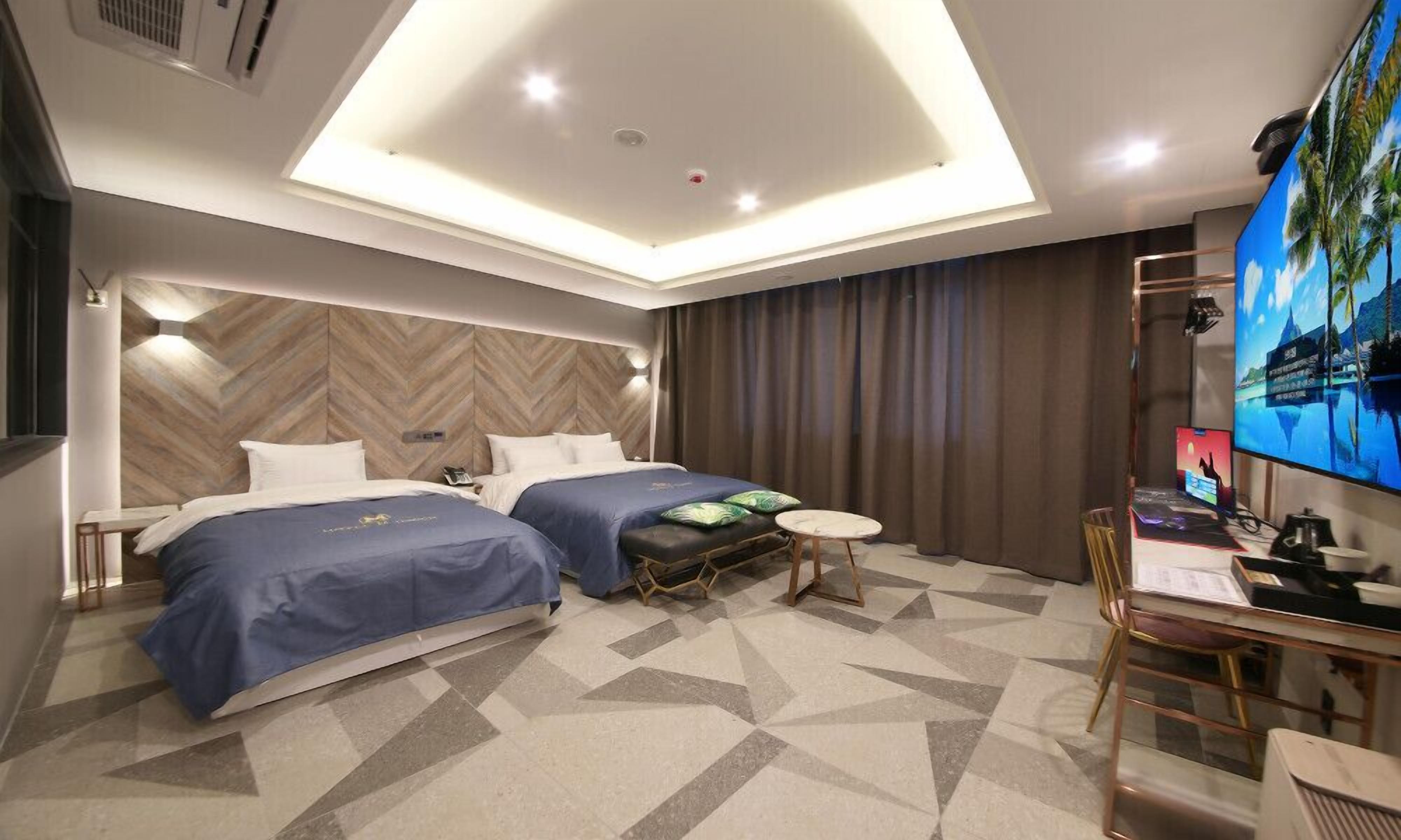 A hotel room with two beds, a desk, a TV, and a modern ceiling design.