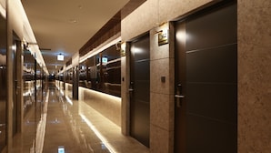 Corredor moderno de hotel com pisos refletivos, portas de elevador escuras e sinalização iluminada.