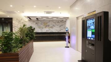 Zona de recepción de un hotel con pared de mármol, mostrador de recepción, exposición de plantas y un quiosco de autoservicio.