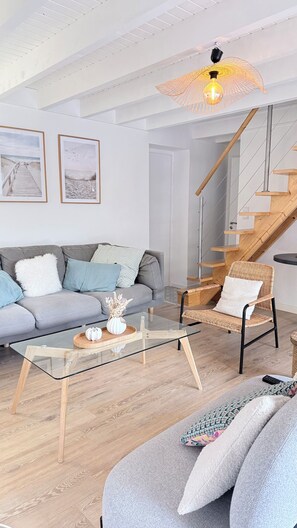 Een moderne woonkamer met een glazen salontafel, een grijze bank, een rieten stoel en een houten trap.