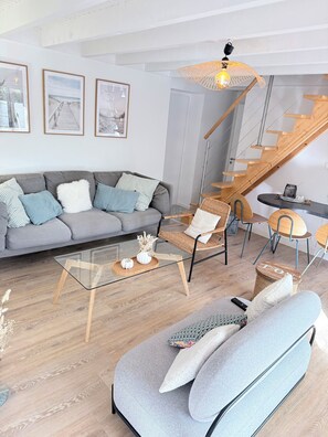 A modern living room with a grey sofa, a glass coffee table, a wicker chair, and a wooden staircase. - Breton house 5 minutes from the beach, with a patio and garden, in Fouesnant – Les Glénan (Fouesnant)