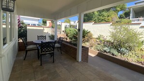 Un patio techado con mesa y sillas, rodeado de un jardín con diversas plantas y un muro de ladrillo.