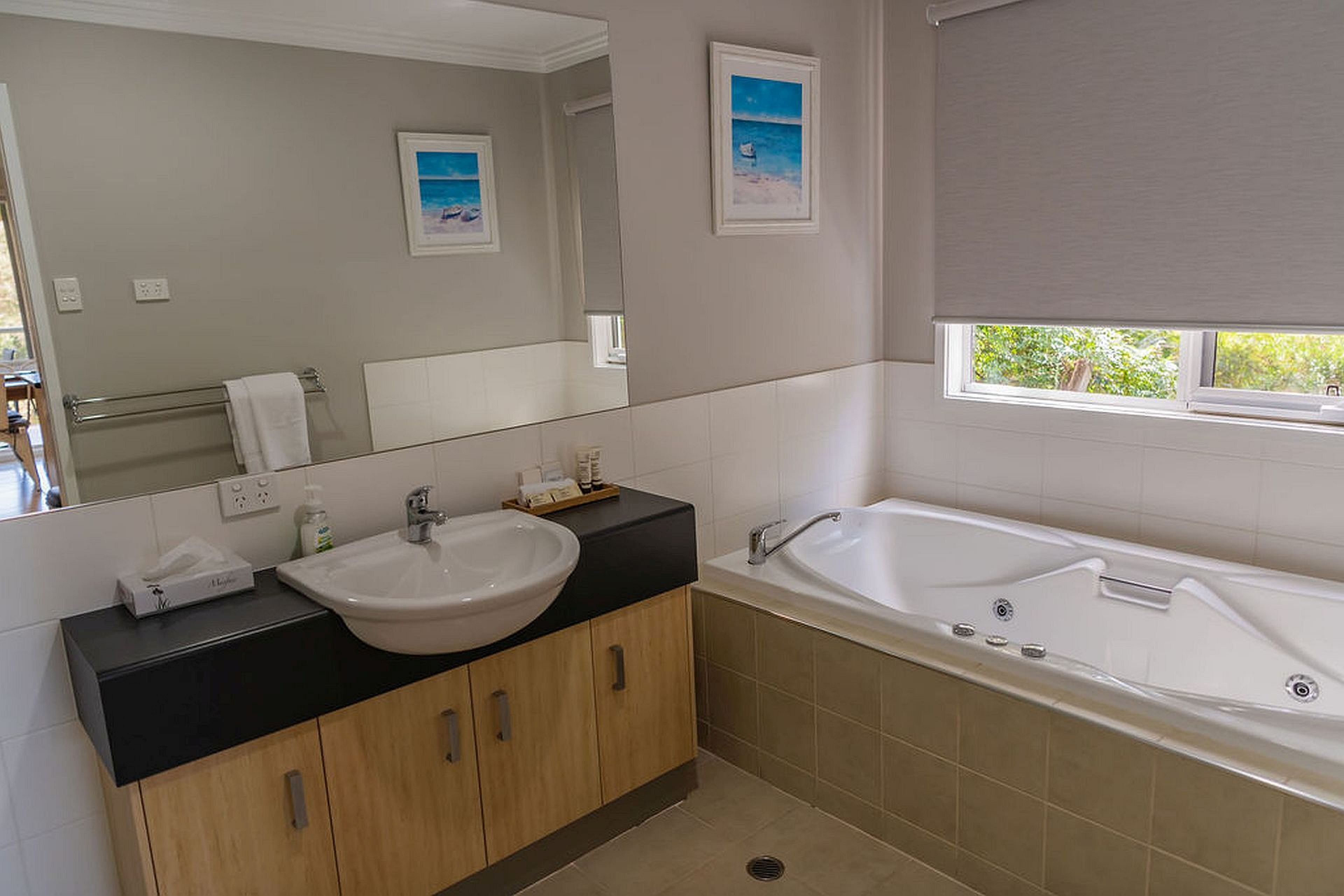 A modern bathroom with a large mirror, a sink, a bathtub, and a window with a view of greenery.