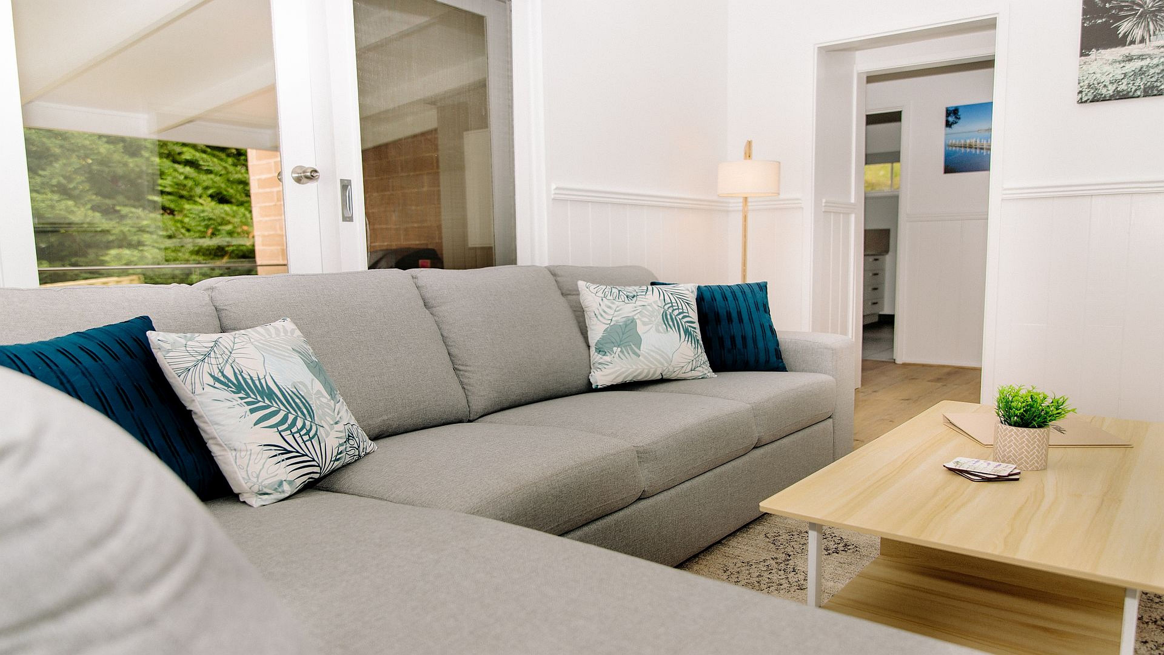 A modern living room with a grey sofa, two patterned cushions, a wooden coffee table, and a view of a garden through a glass door.
