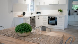 A modern kitchen with white cabinets, a wooden table with a woven placemat, two wine glasses, and a potted plant. - McMillans of Metung Coastal Resort (Metung)