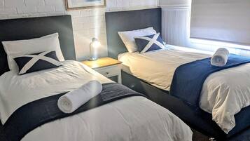 Two adjacent beds with white linens, dark headboards, and a small bedside table with a lamp.
