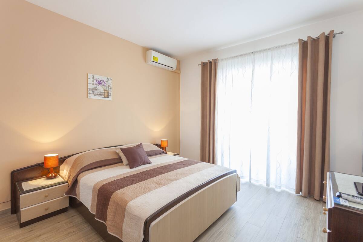 A neatly made bed with a striped blanket, two bedside lamps, a wall-mounted air conditioner, and a window with curtains.