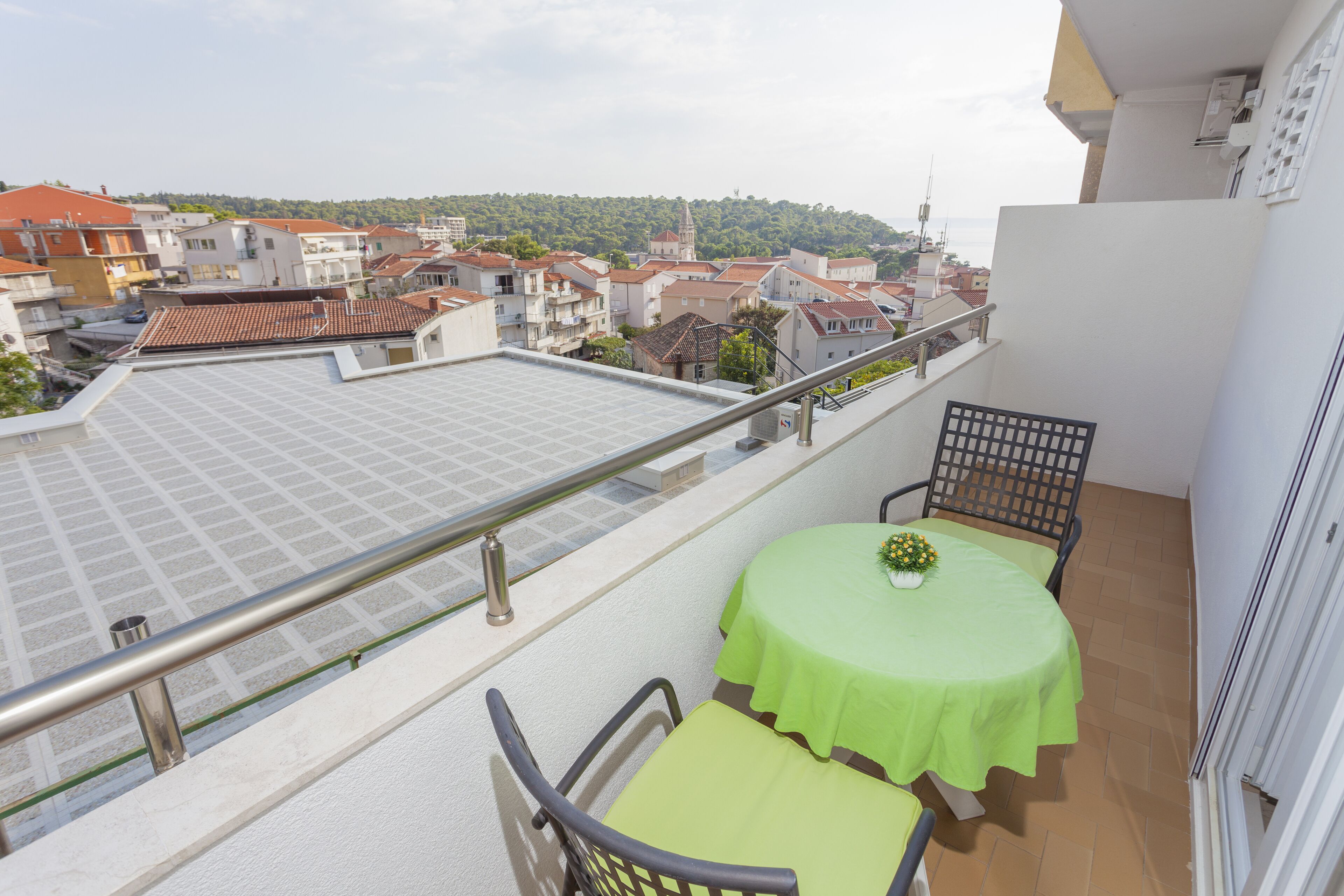 Uma varanda com mesa e cadeiras, com vista para um horizonte urbano com edifícios de telhado vermelho e uma colina ao longe.