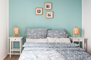 A bedroom with teal walls, a bed with patterned bedding, two bedside tables with wicker lamps, and three framed pictures on the wall.