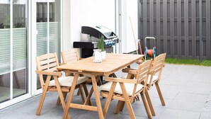 Une table et des chaises en bois sont disposées sur une terrasse, avec un barbecue en arrière-plan.
