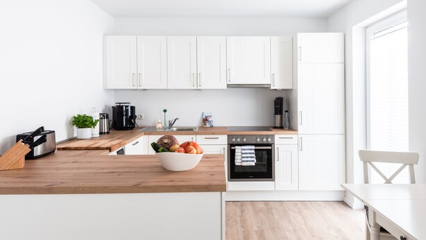 Une cuisine moderne avec des meubles blancs, un plan de travail en bois, un four noir et un coin repas comprenant une table et des chaises blanches.