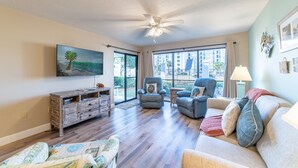 A living room with a ceiling fan, a large window, a sofa, a chair, and a wooden cabinet. - SanDBeach Villa, Edgewater Villa 3302-1st Floor-2 Patios-2 Bd/2Ba-Free Sandy Beach Tram! (Panama City Beach)