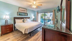 A bedroom with a bed, bedside tables, a ceiling fan, a window with curtains, and a view of buildings outside. - SanDBeach Villa, Edgewater Villa 3302-1st Floor-2 Patios-2 Bd/2Ba-Free Sandy Beach Tram! (Panama City Beach)
