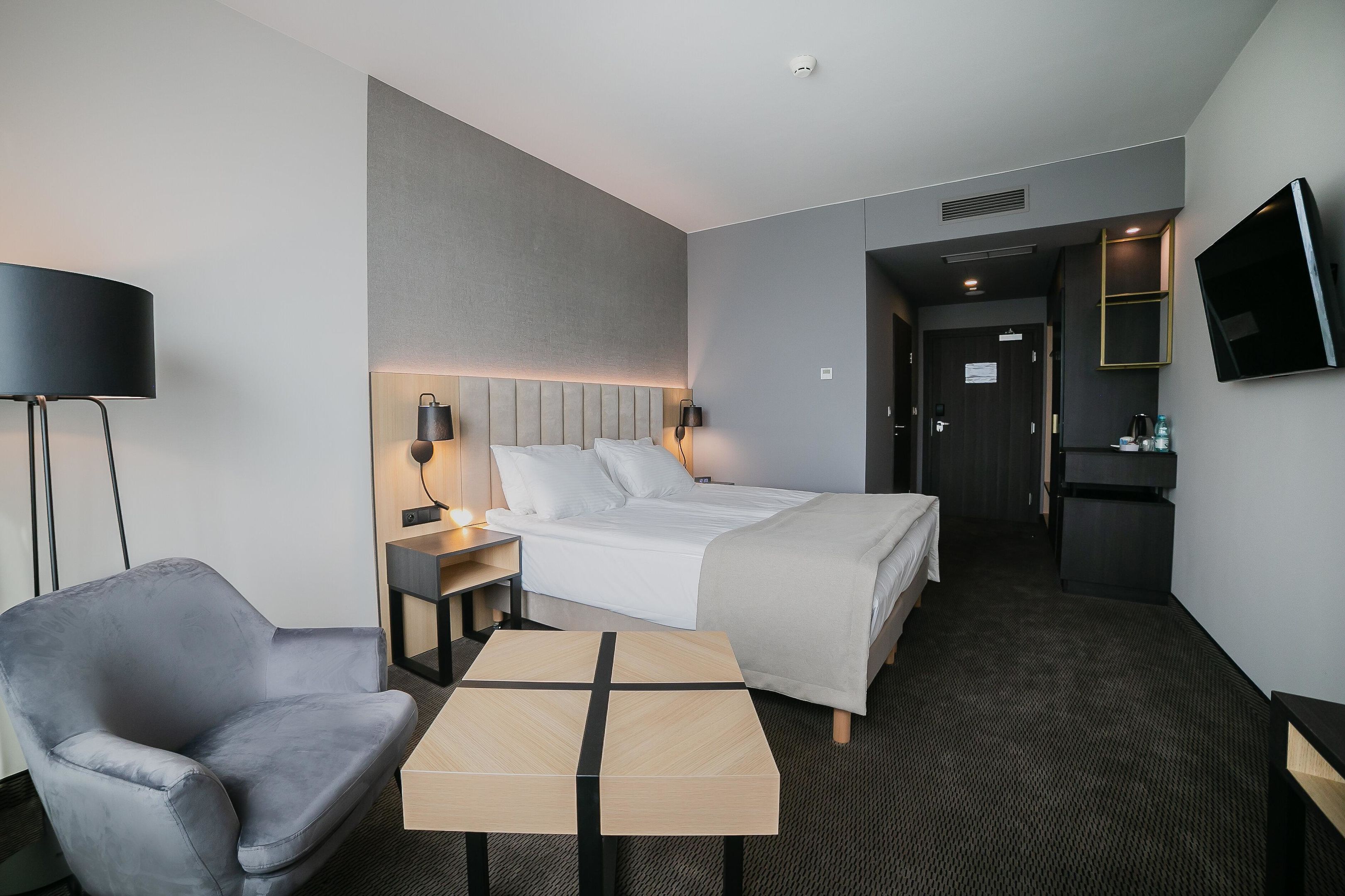 Une chambre d’hôtel moderne avec un lit, un fauteuil gris, une petite table, une lampe de chevet et un téléviseur fixé au mur.