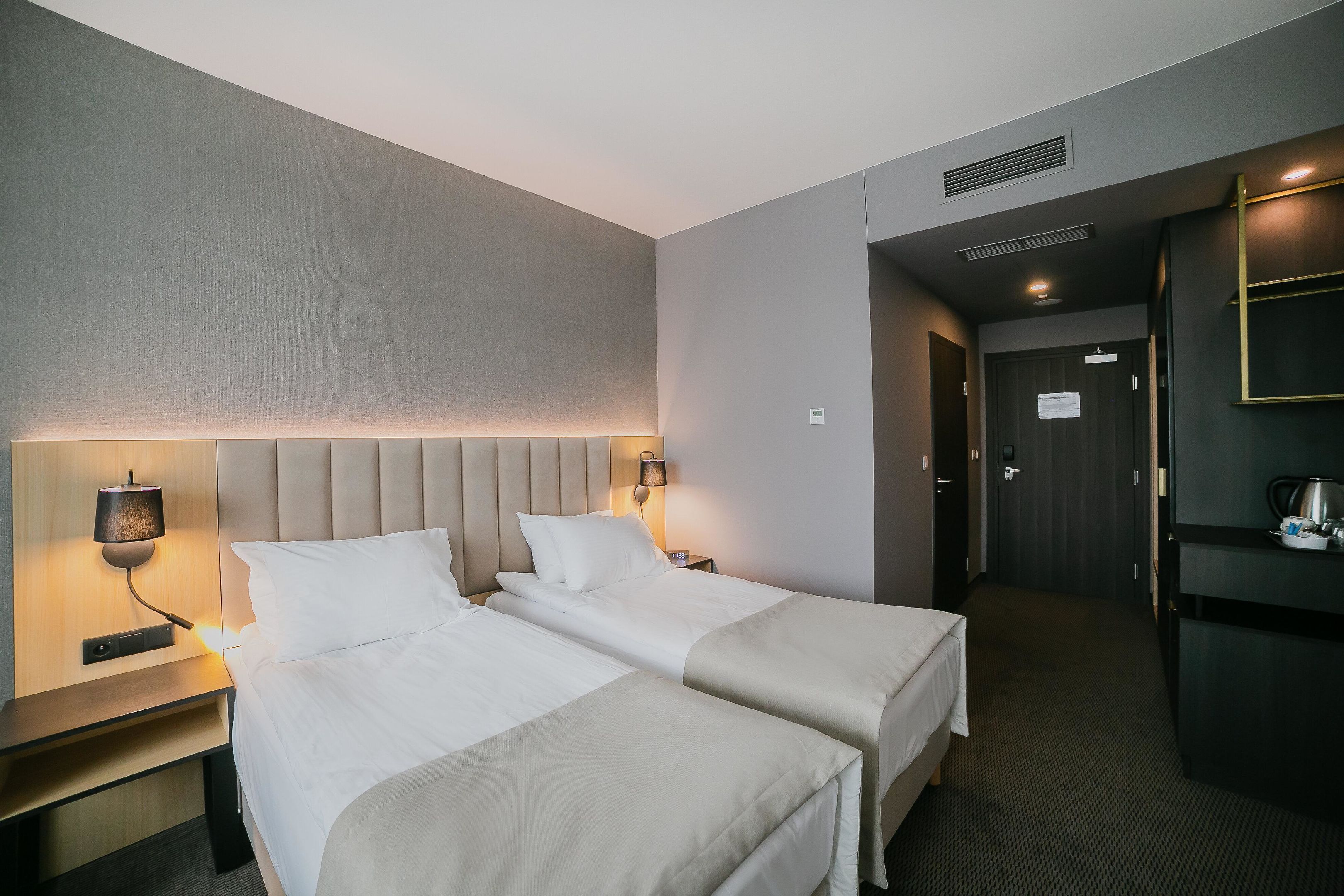 A hotel room with two beds, a bedside table with a lamp, a small table with a kettle, and a dark-colored wardrobe.