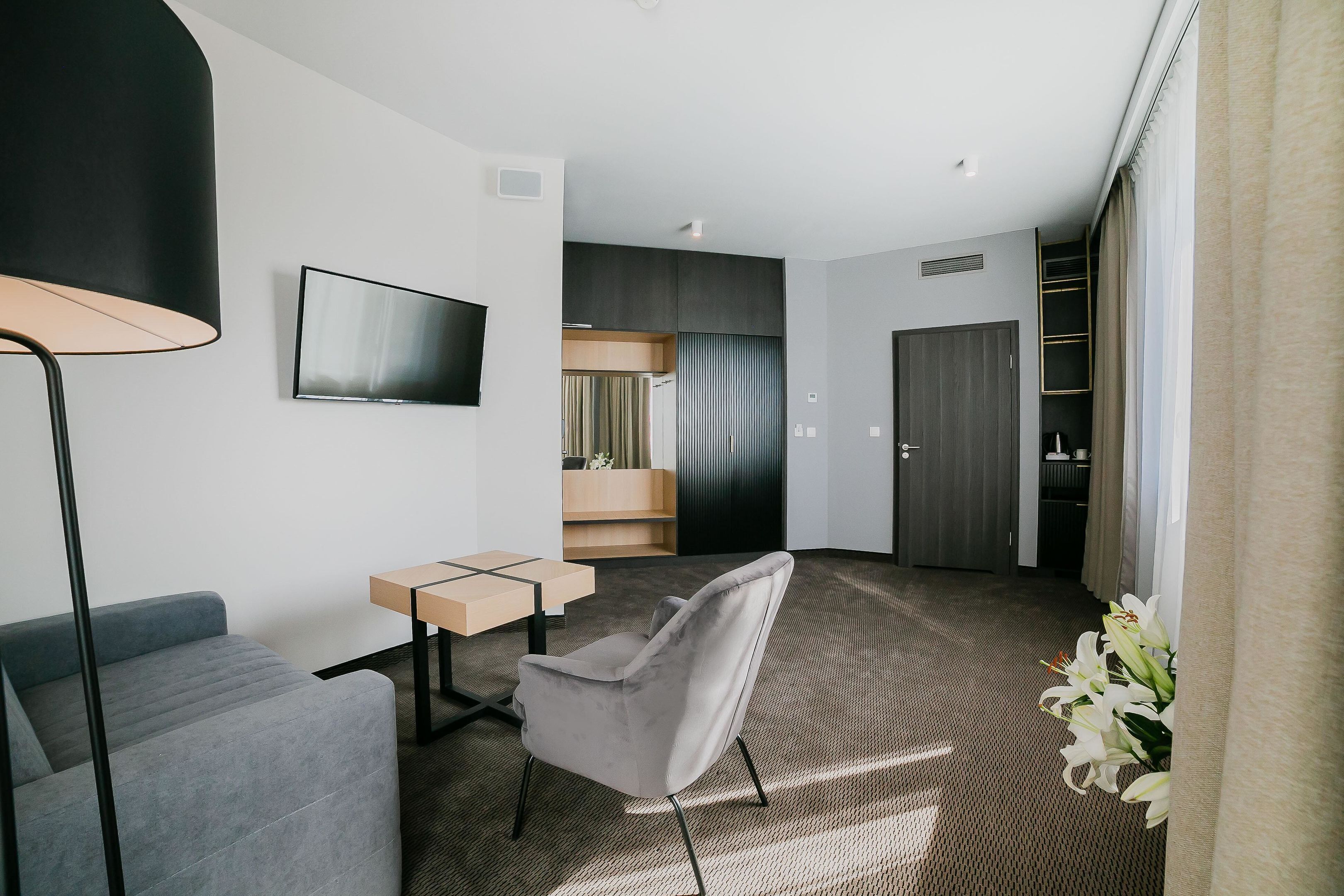 Una habitación de hotel moderna con un sofá gris, una mesita, un sillón gris, un televisor montado en la pared, un armario y una escalera que lleva al piso superior.