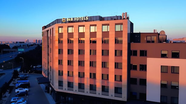 A multi-story hotel with a prominent 'B&B HOTELS' sign, a parking lot with several cars, and a cityscape in the background. - B&B HOTEL Warsaw East (Warsaw)