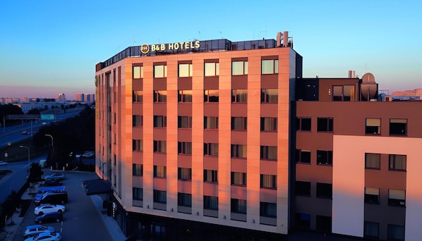 A multi-story hotel with a prominent 'B&B HOTELS' sign, a parking lot with several cars, and a cityscape in the background. - B&B HOTEL Warsaw East (Warsaw)