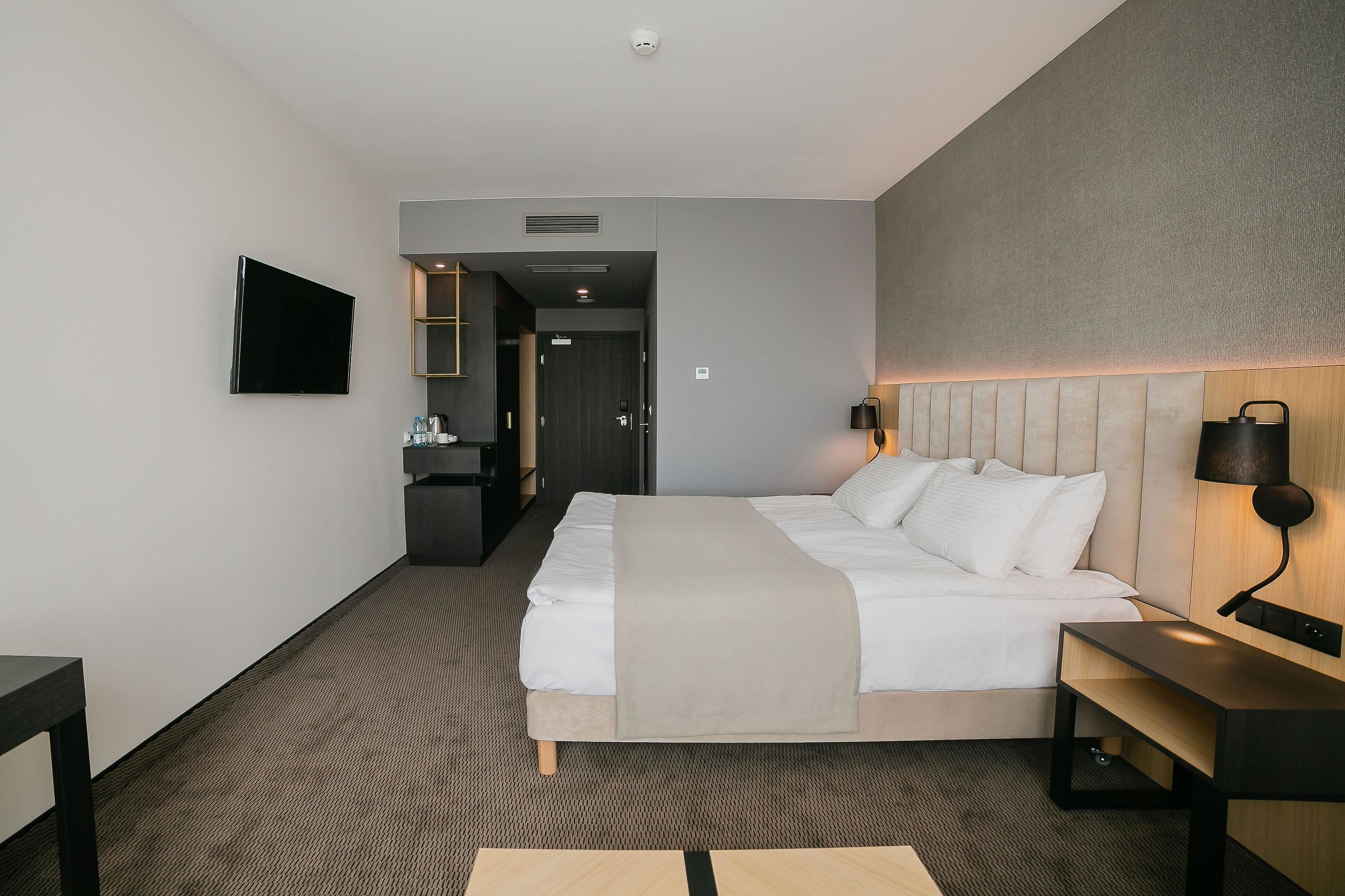 A modern hotel room with a large bed, bedside tables, a flat-screen TV, and a headboard with integrated lighting.