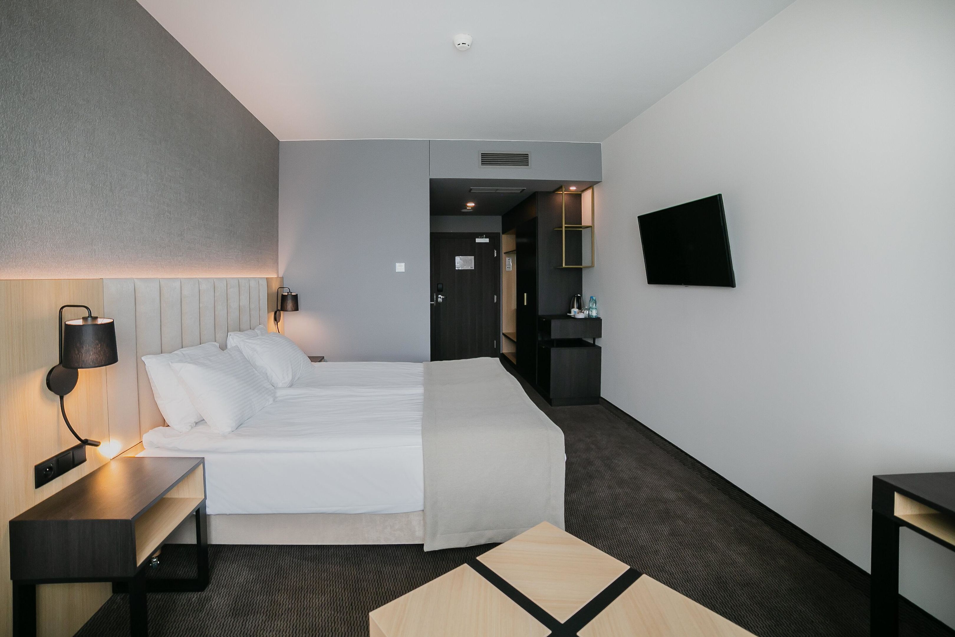 Una habitación de hotel moderna con una cama grande, mesita de noche, lámpara y televisor de pantalla plana montado en la pared.