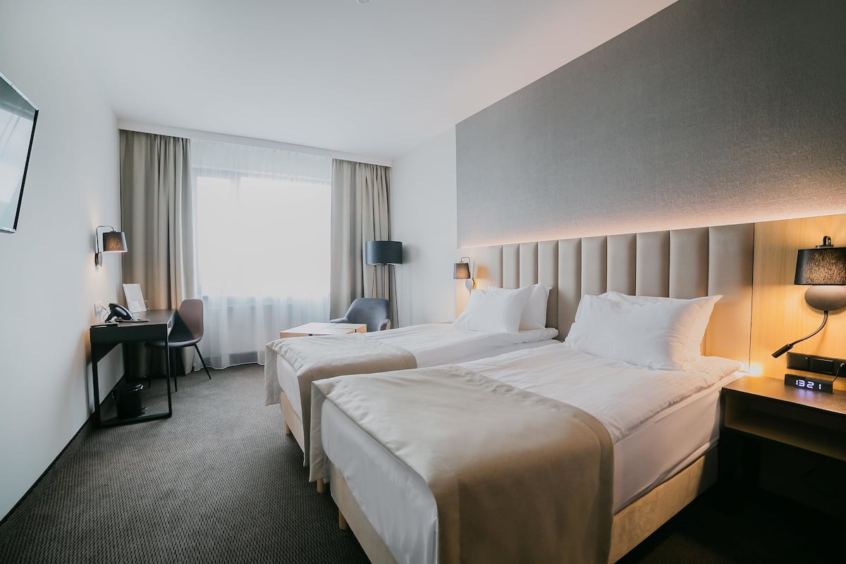 Hotellihuone, jossa on kaksi sänkyä, työpöytä, tuoli, lamppu ja yöpöydällä oleva kello.