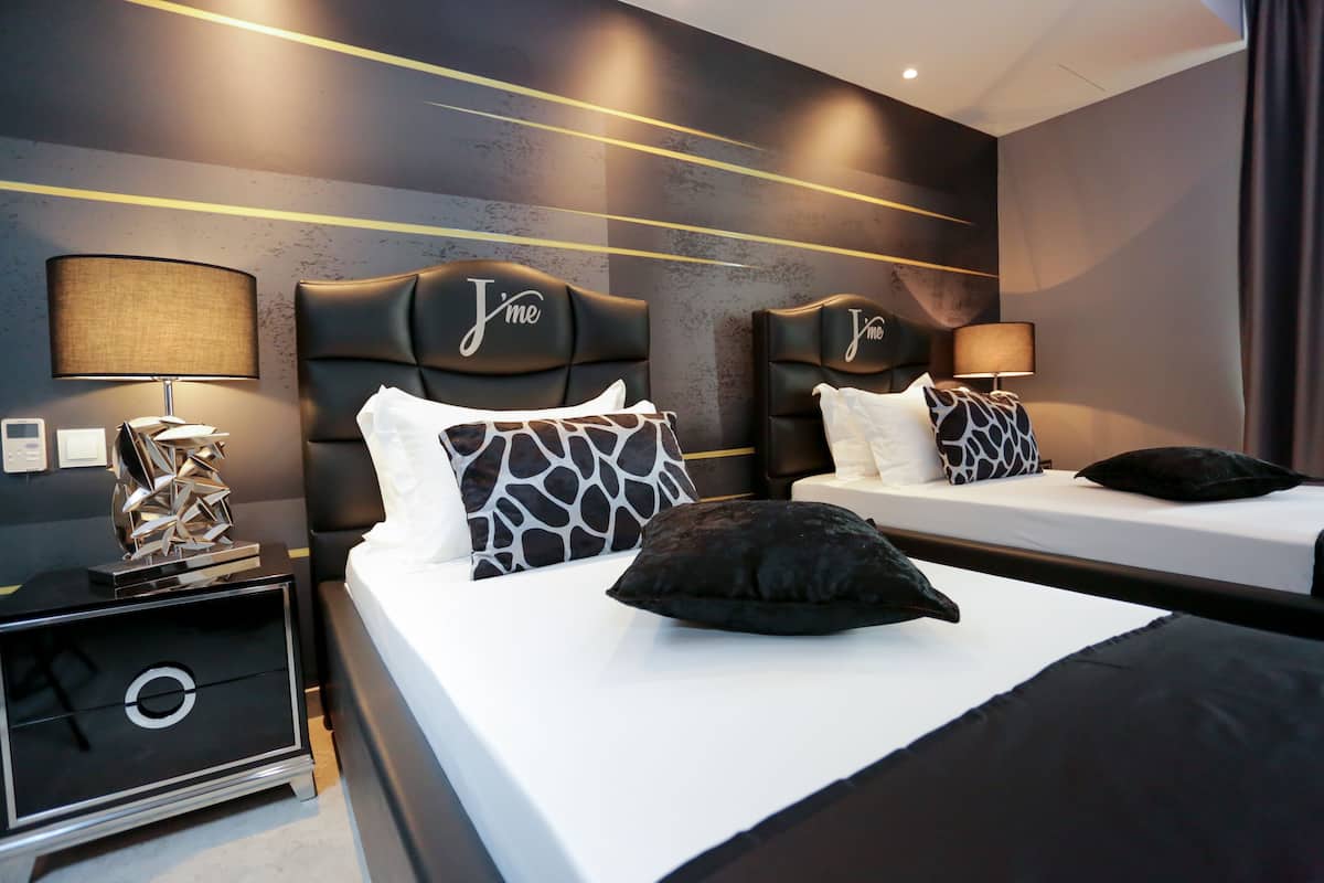 Ein Hotelzimmer mit zwei Betten, jeweils mit einem Kopfteil mit dem Schriftzug 'Jime'. An beiden Seiten der Betten befinden sich Lampen, Nachttische mit einer Schublade sowie ein dekoratives Element auf dem Nachttisch.