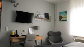 A hotel room with a bed, a desk with a chair, a television, a lamp, and a shelf with items.
