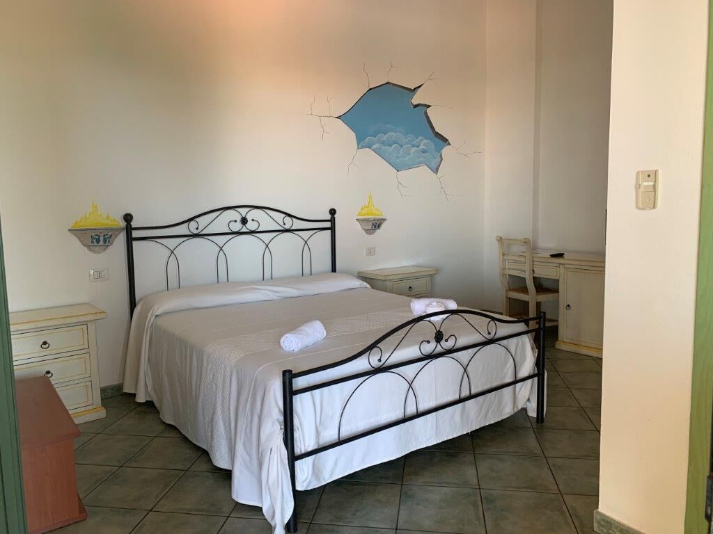 Un dormitorio con estructura de cama de metal, ropa de cama blanca, dos mesitas de noche con lámparas y una pared con diseño agrietado.