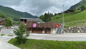 A building with a sign for 'La Grande Maison' and a 'Pizza Pie' sign, surrounded by greenery and mountains. - Charming Studio, Ski-In/Out, Sleeps 4, Balcony View, Parking, Near Village Center (Le Grand-Bornand)