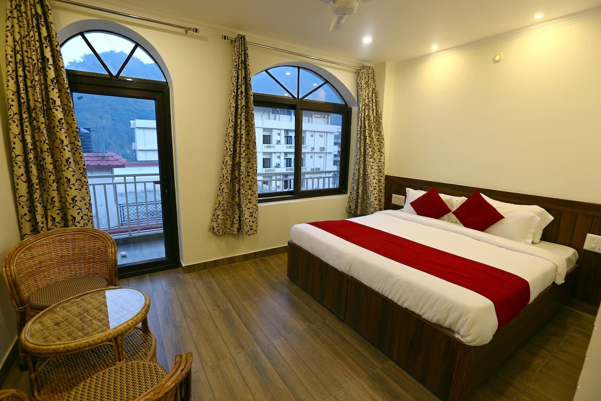 A hotel room with a large bed, two windows with curtains, a wicker chair, and a balcony with a railing.