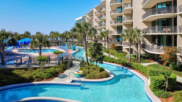 Un resort con una gran piscina, palmeras y un edificio de varios pisos con balcones.