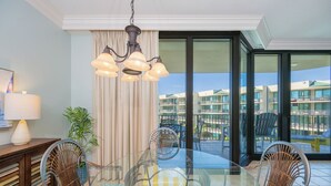 Un comedor con una mesa de vidrio y sillas de mimbre, un candelabro y vistas a un edificio a través de grandes ventanales.