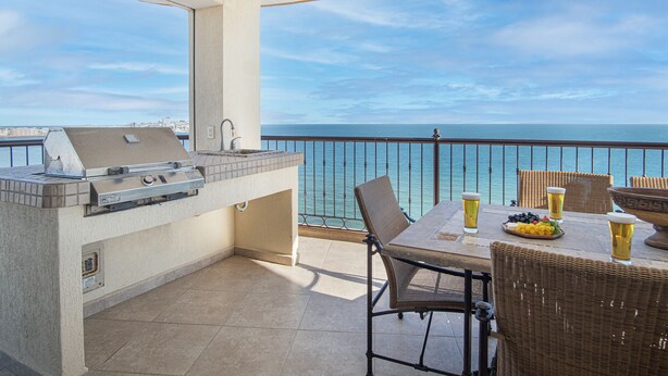 Een balkon met een barbecue, eettafel, stoelen en uitzicht op zee.